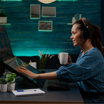 A girl sitting in a blue tone home office, smiling, holding a white mug and touching a double monitor on the desk, with a mouse, notes, pen, and flowers also on the desk.